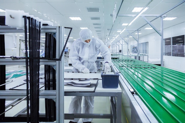 Trabajadores de fábrica con trajes de laboratorio blancos y máscaras faciales que producen televisores en una línea de montaje verde con algunos equipos modernos