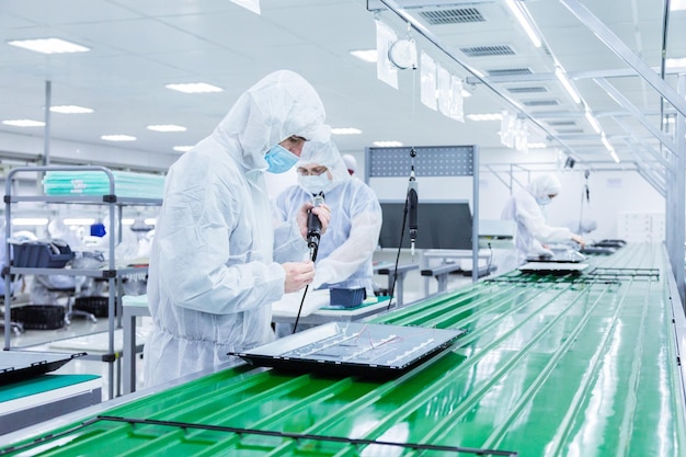 Trabajadores de fábrica con trajes de laboratorio blancos y máscaras faciales que producen televisores en una línea de montaje verde con algunos equipos modernos