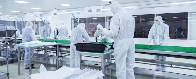 Trabajadores de fábrica con trajes de laboratorio blancos y máscaras faciales que producen televisores en una línea de montaje verde con algunos equipos modernos
