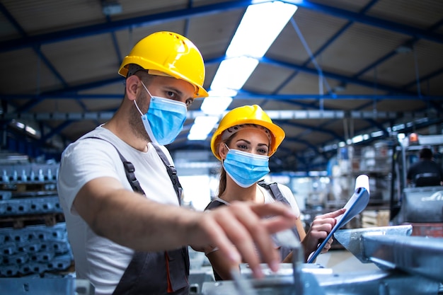 Trabajadores industriales con mascarillas protegidas contra el virus corona discutiendo sobre piezas metálicas en la fábrica