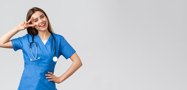 Trabajadores médicos cuidado de la salud covid19 y concepto de vacunación Enfermera o doctora atractiva alegre con estetoscopio de matorrales azules muestra el signo de la paz manténgase optimista de pie fondo gris