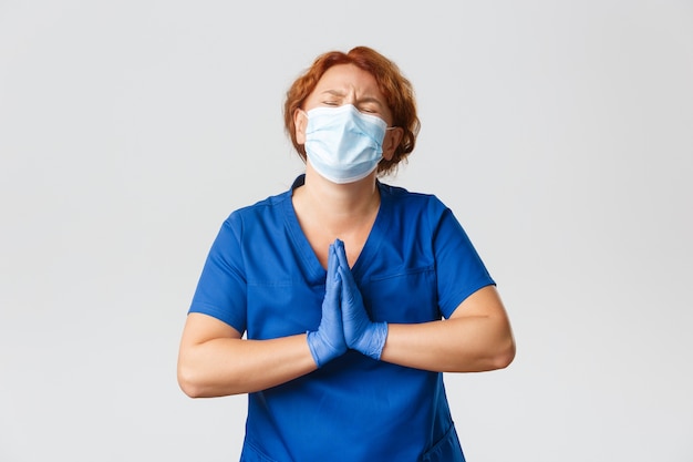 Trabajadores médicos, pandemia, concepto de coronavirus. Doctora pelirroja mendigando angustiada en mascarilla y guantes de goma suplicando, suplicando, pidiendo ayuda, pared gris.