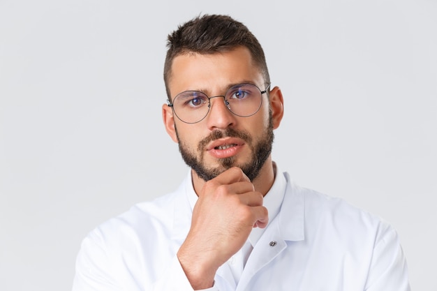 Trabajadores de la salud, coronavirus, concepto de pandemia covid-19. Primer plano de un médico profesional de aspecto serio con gafas y bata blanca, toque la barbilla pensativo, pensando, tomando decisiones sobre el paciente