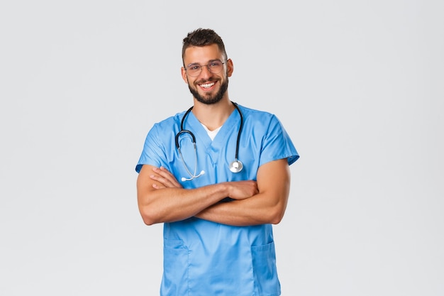 Trabajadores de la salud, medicina, covid-19 y concepto de autocuarentena pandémica. Doctor atractivo sonriente en matorrales y gafas, estetoscopio sobre el cuello, pecho de brazos cruzados, listo para ayudar a los pacientes