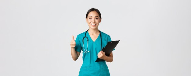 Trabajadores de la salud, prevención de virus, concepto de campaña de cuarentena. Una doctora bastante asiática sonriente, una pasante en batas muestra el pulgar hacia arriba, sostiene el portapapeles, trae resultados positivos, fondo blanco.
