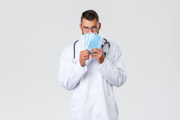 Trabajadores de la salud, seguro médico, concepto de pandemia y covid-19. Doctor guapo divertido, médico hispano con bata blanca y gafas, mostrando máscaras médicas, úsela durante el coronavirus