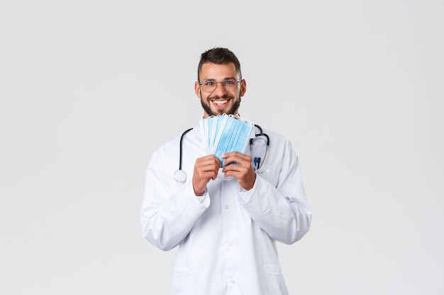 Trabajadores de la salud, seguro médico, concepto de pandemia y covid-19. Feliz y guapo médico con gafas y batas, mostrando máscaras médicas y sonriendo, recomienda protegerse durante la corona.