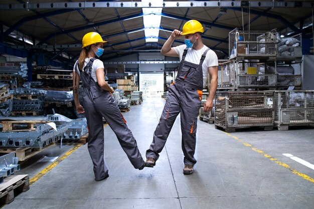 Trabajadores vestidos con uniformes y casco en la fábrica tocándose con las piernas y saludando debido al virus de la corona y la infección