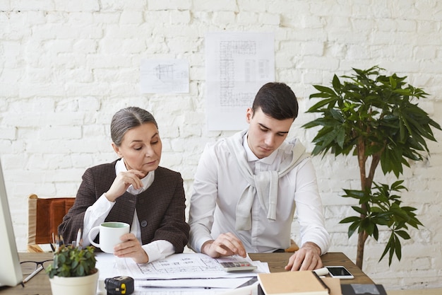 Trabajando en equipo. Arquitecto masculino joven seriamente afeitado y enfocado haciendo cálculos usando la calculadora sentado en la oficina con su jefa de cabello gris, estudiando dibujos frente a ellos