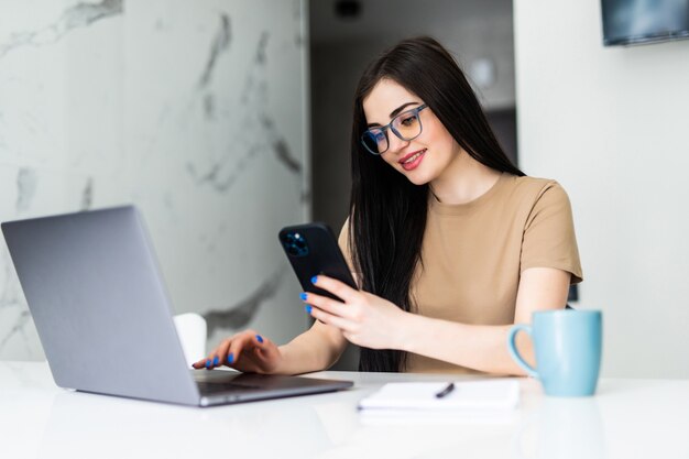 Trabajar en casa. Mujer joven usa una computadora portátil para trabajar en la cocina y usa el teléfono