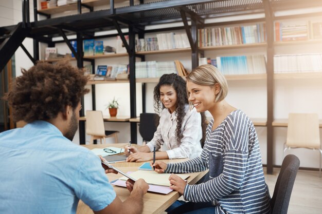 Trabajo colaborativo. Grupo de gerentes de proyectos jóvenes que trabajan en nuevas startups, planes de análisis. Tres perspectivas profesionales jóvenes sentados en la biblioteca moderna en reunión.