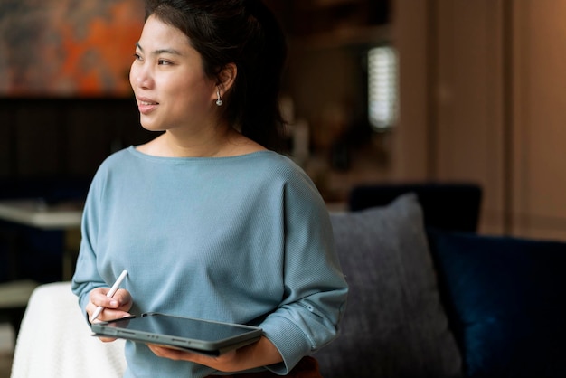 Trabajo informal en casa, relájese informal usando un dispositivo de tableta en la sala de estar, una mujer adulta asiática, escribiendo a mano, busque o envíe mensajes de texto a través del sofá de una computadora portátil, trabajando desde casa con felicidad, concepto de ideas sonrientes pacíficas