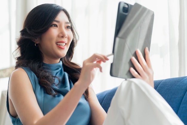 Trabajo informal en casa, relájese informal usando un dispositivo de tableta en la sala de estar, una mujer adulta asiática, escribiendo a mano, busque o envíe mensajes de texto a través del sofá de una computadora portátil, trabajando desde casa con felicidad, concepto de ideas sonrientes pacíficas