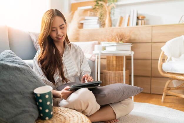 Trabajo informal en casa, relájese informal usando un dispositivo de tableta en la sala de estar, una mujer adulta asiática, escribiendo a mano, busque o envíe mensajes de texto a través del sofá de una computadora portátil, trabajando desde casa con felicidad, concepto de ideas sonrientes pacíficas