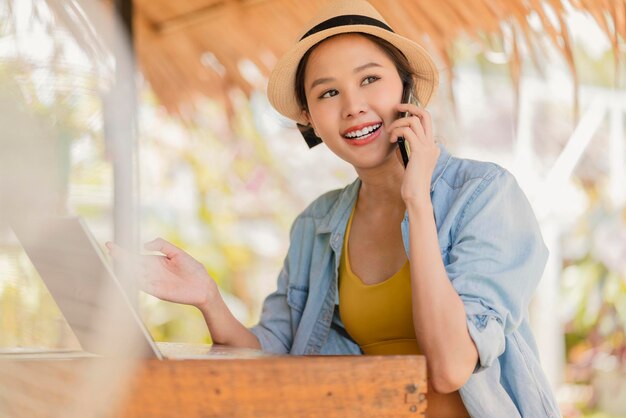 Trabajo y vacaciones mujer asiática nómada digital estilo de vida milenario mano sostener conversación de teléfono inteligente con el cliente en el extranjero reunión a distanciamujer asiática ropa casual caminando hablando en la playa océano