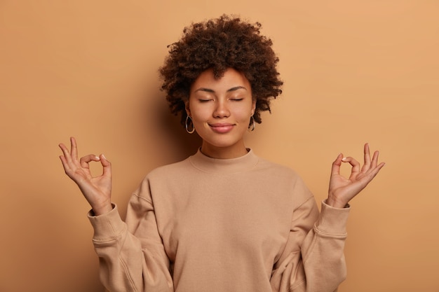 Tranquila y aliviada mujer de piel oscura respira hondo, mantiene las manos a los lados en gesto zen, alcanza el nirvana y practica yoga, se para con los ojos cerrados, se para sin estrés contra la pared marrón