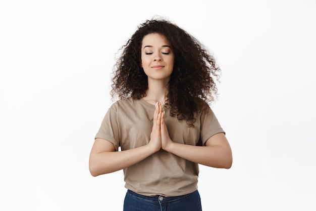 Tranquila joven meditando, muestra el gesto de rezar namaste, cierra los ojos y sonríe, pide deseos, suplica a Dios, de pie sobre fondo blanco