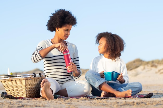 Tranquila madre afroamericana e hija en un picnic en la playa. Mujeres con ropa informal sentadas en una manta, sosteniendo termos y tazas. Familia, relajación, concepto de naturaleza.