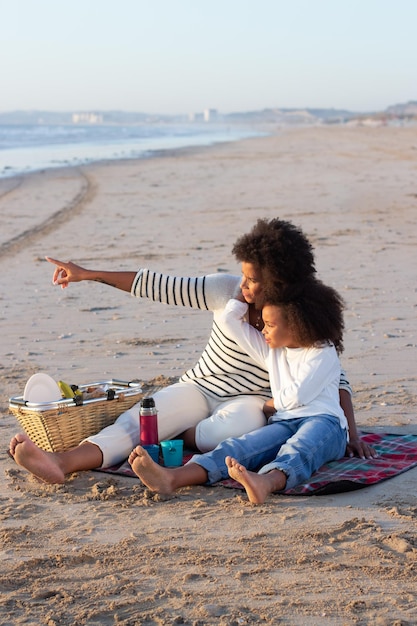 Tranquila madre e hija haciendo un picnic en la playa. Madre e hija afroamericanas con ropa informal sentadas en una manta, discutiendo la puesta de sol. Familia, relajación, concepto de naturaleza.
