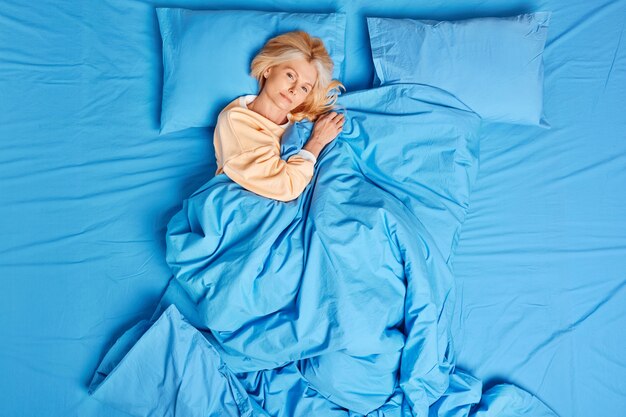 Tranquila mujer europea de mediana edad se despierta satisfecha después de ver buenos sueños posa bien dormida bajo una manta azul viste pijama se siente cómoda disfruta de un día de descanso. Hora de dormir y concepto acogedor de la mañana.