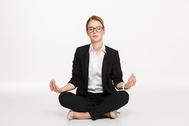 Tranquila rubia mujer de negocios en anteojos meditación en estudio con los ojos cerrados sobre la pared blanca