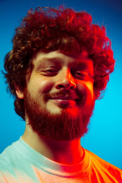 Tranquilo, encantado. Retrato de hombre caucásico de cerca aislado en la pared azul en luz de neón roja. Hermoso modelo masculino, pelo rizado rojo. Concepto de emociones humanas, expresión facial, ventas, publicidad.