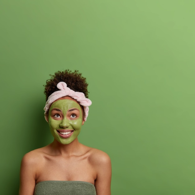 Tratamiento de belleza spa y concepto de cuidado de la piel. La mujer positiva se aplica una máscara de peeling facial, se mantiene joven y hermosa concentrada arriba con amplias sonrisas, usa una diadema, posa sobre el espacio de copia de la pared verde