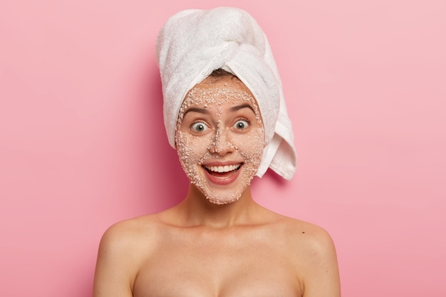 Foto gratuita tratamiento facial. hermosa mujer feliz con una sonrisa encantadora, elimina toxinas y puntos negros en la cara, aplica exfoliante natural de gránulos de sal marina blanca, saca zuecos, tiene un cuerpo desnudo y bien cuidado.