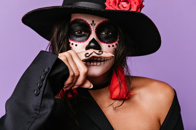 La traviesa chica de ojos marrones con caras artísticas se divierte poniéndose bigotes pintados en la boca. Foto de mujer elegante en traje negro con detalles en rojo.