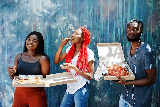 Tres alegres amigos afroamericanos sosteniendo cajas de pizza contra la pared de pintura