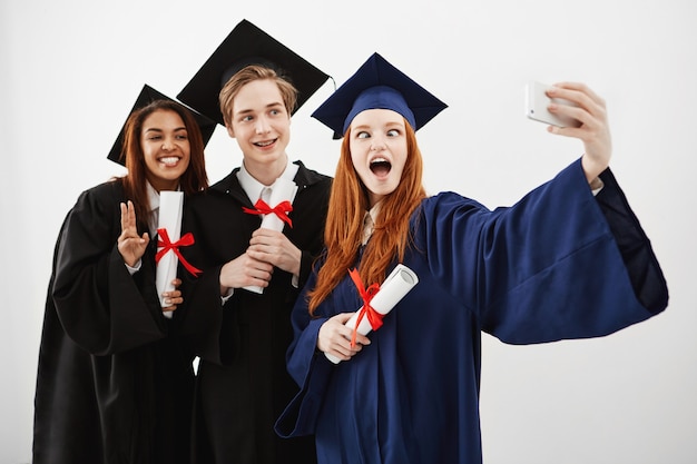 Tres alegres graduados felices jugando y divirtiéndose sonriendo haciendo selfie con diplomas en las manos, futuros abogados.