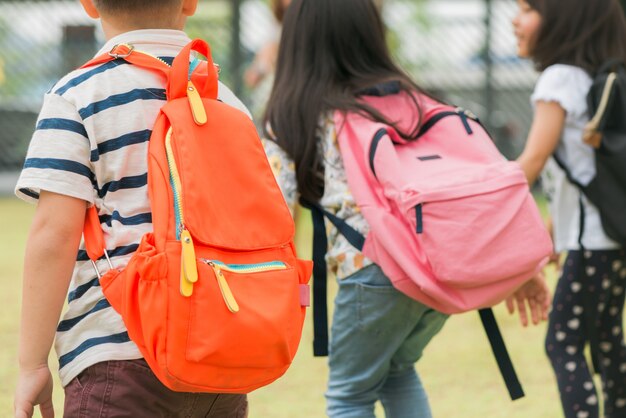 Tres alumnos de la escuela primaria van de la mano. Muchacho y muchacha con los bolsos de escuela detrás de la parte posterior. Comienzo de las clases. Día cálido de otoño. De vuelta a la escuela. Pequeños estudiantes de primer grado.