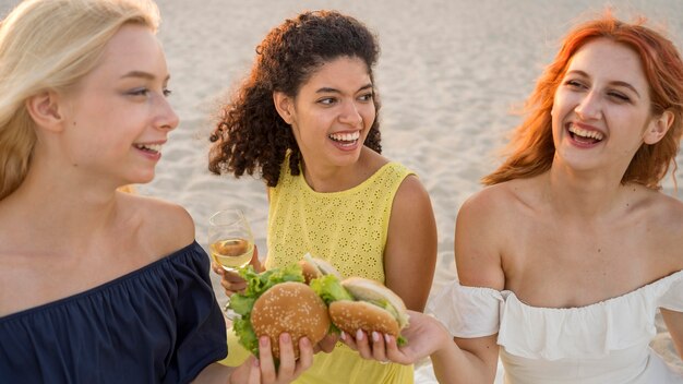 Tres amigas sonrientes disfrutando de hamburguesas juntos en la playa