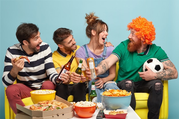 Tres amigos felices miran al hombre barbudo divertido con peluca, tintinean botellas de cerveza, comen pizza, se divierten mientras miran un partido de fútbol en la televisión