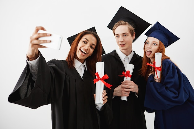 Tres amigos graduados internacionales se regocijan en mantos haciendo una selfie en un teléfono. Futuros especialistas o médicos divirtiéndose con sus diplomas sobre pared blanca.