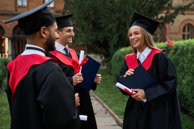 Tres amigos graduados sonrientes en batas de graduación hablando en el campus con diploma.