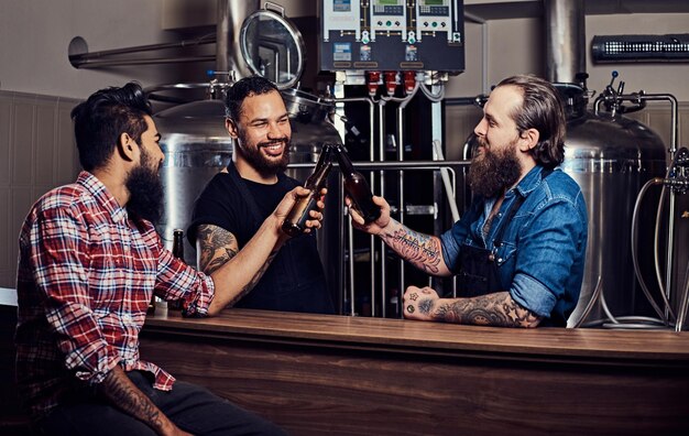 Tres amigos interraciales barbudos beben cerveza artesanal y hablan en una cervecería. Dos trabajadores hipster en delantales beben cerveza con su amigo en una fábrica de cerveza. Viejos amigos se reunían para beber cerveza y charlar.