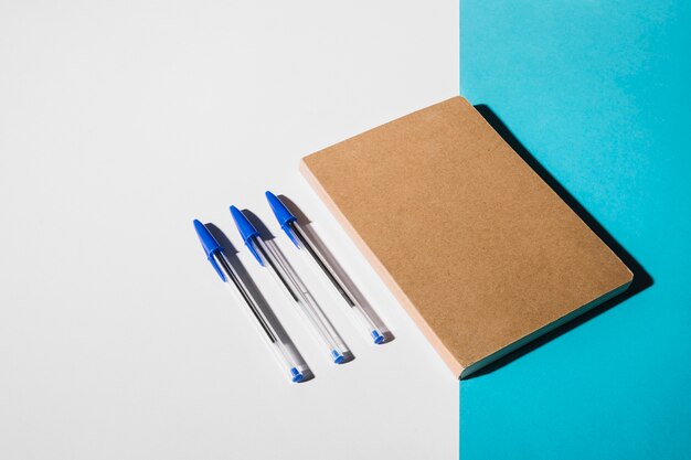Tres bolígrafos y libro cerrado sobre doble fondo blanco y azul