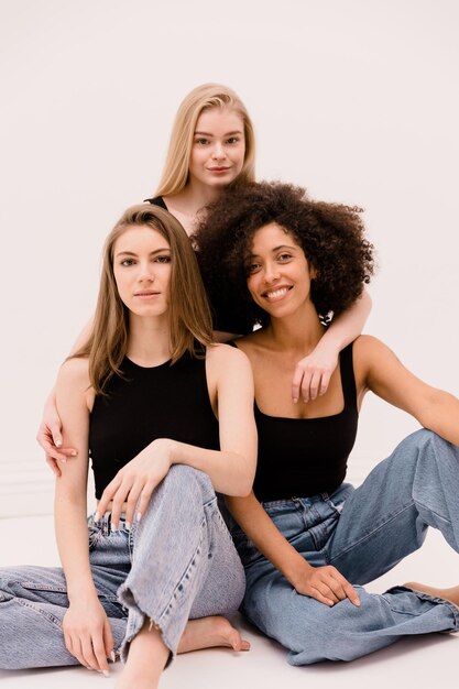 Tres bonitas mujeres interraciales jóvenes de cabello claro y oscuro en tops negros y jeans miran a la cámara sobre fondo blanco