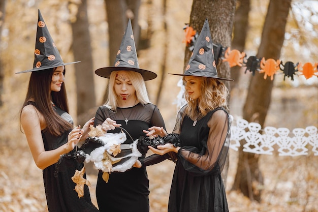 Tres brujas de chicas jóvenes en el bosque en Halloween. Chicas con vestidos negros y sombrero de cono. Bruja sosteniendo un caldero de mago.