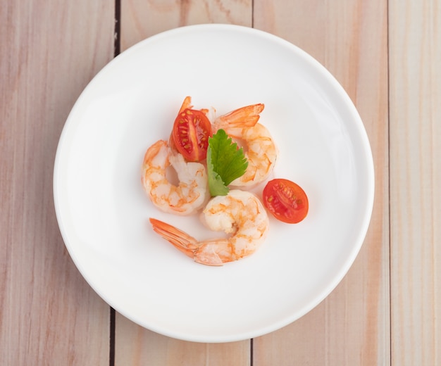 Tres camarones frescos y medio tomate en un plato blanco sobre una madera.