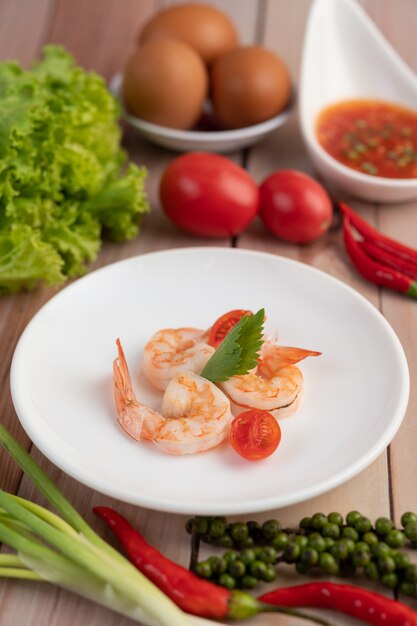 Tres camarones frescos y medio tomate en un plato blanco sobre una madera.