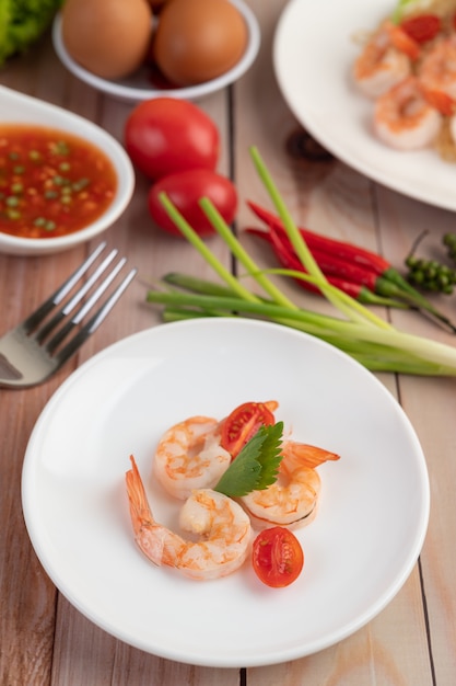 Tres camarones frescos y medio tomate en un plato blanco sobre una madera.