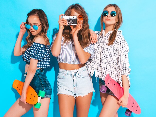 Tres chicas guapas con estilo hermosas sonrientes con patinetas coloridas centavo Mujeres en ropa de camisa a cuadros de verano posando. Modelos tomando fotos con la cámara de fotos retro