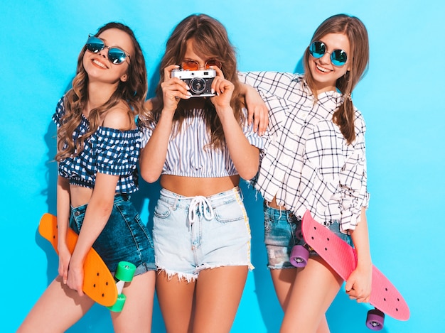 Tres chicas guapas con estilo hermosas sonrientes con patinetas coloridas centavo Mujeres en ropa de camisa a cuadros de verano posando. Modelos tomando fotos con la cámara de fotos retro