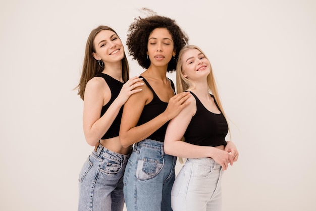 Tres chicas jóvenes interraciales en tops negros y jeans se paran una por una sobre fondo blanco