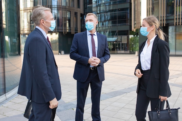 Tres colegas de negocios profesionales en máscaras faciales discutiendo el trato afuera. Gerentes de contenido exitosos de pie en la calle y hablando sobre el trabajo. Concepto de negociación, protección y asociación