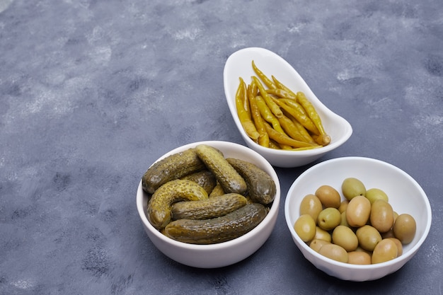 Tres cuencos de pepinos encurtidos, pimientos y aceitunas sobre fondo azul.