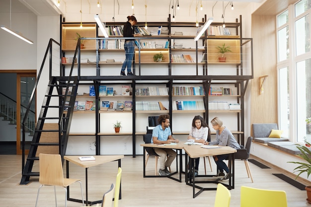 Foto gratuita tres diseñadores entusiastas felices discutiendo ideas de negocios para el próximo proyecto sentados a la mesa con papeles en la biblioteca moderna y brillante confort trabajo en equipo con amigos. startup, concepto de negocio