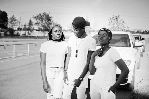 Tres elegantes amigos afroamericanos visten ropa blanca contra dos autos de lujo Moda callejera de jóvenes negros Hombre negro con dos niñas africanas